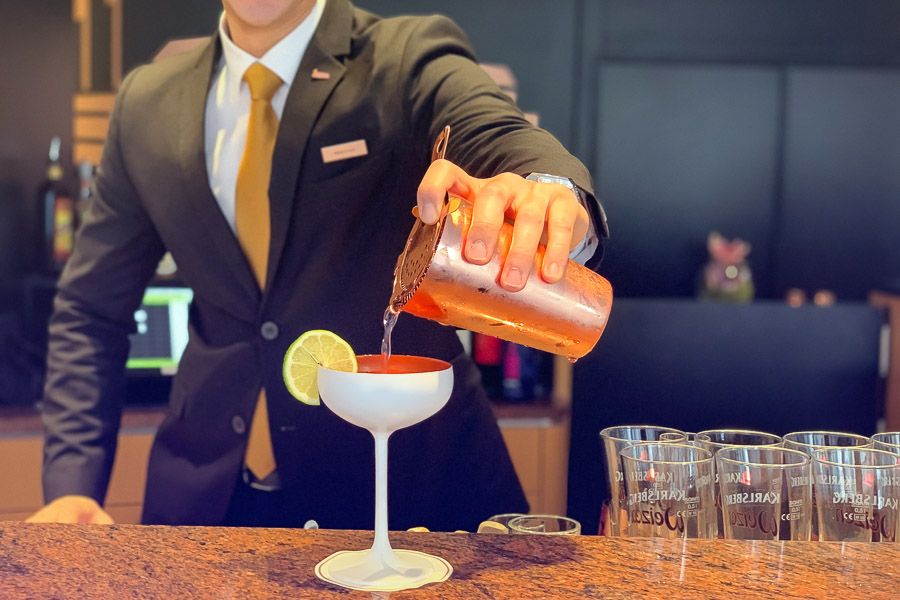 A bartender in a suit pours a cocktail from a copper shaker into a white coupe with a slice of lime
