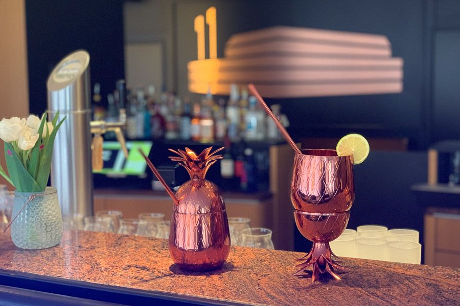 Zwei Cocktails in kupfernen Ananas- und Kelchbechern auf dem Bartresen, dahinter Flaschenregal und Leuchtlogo