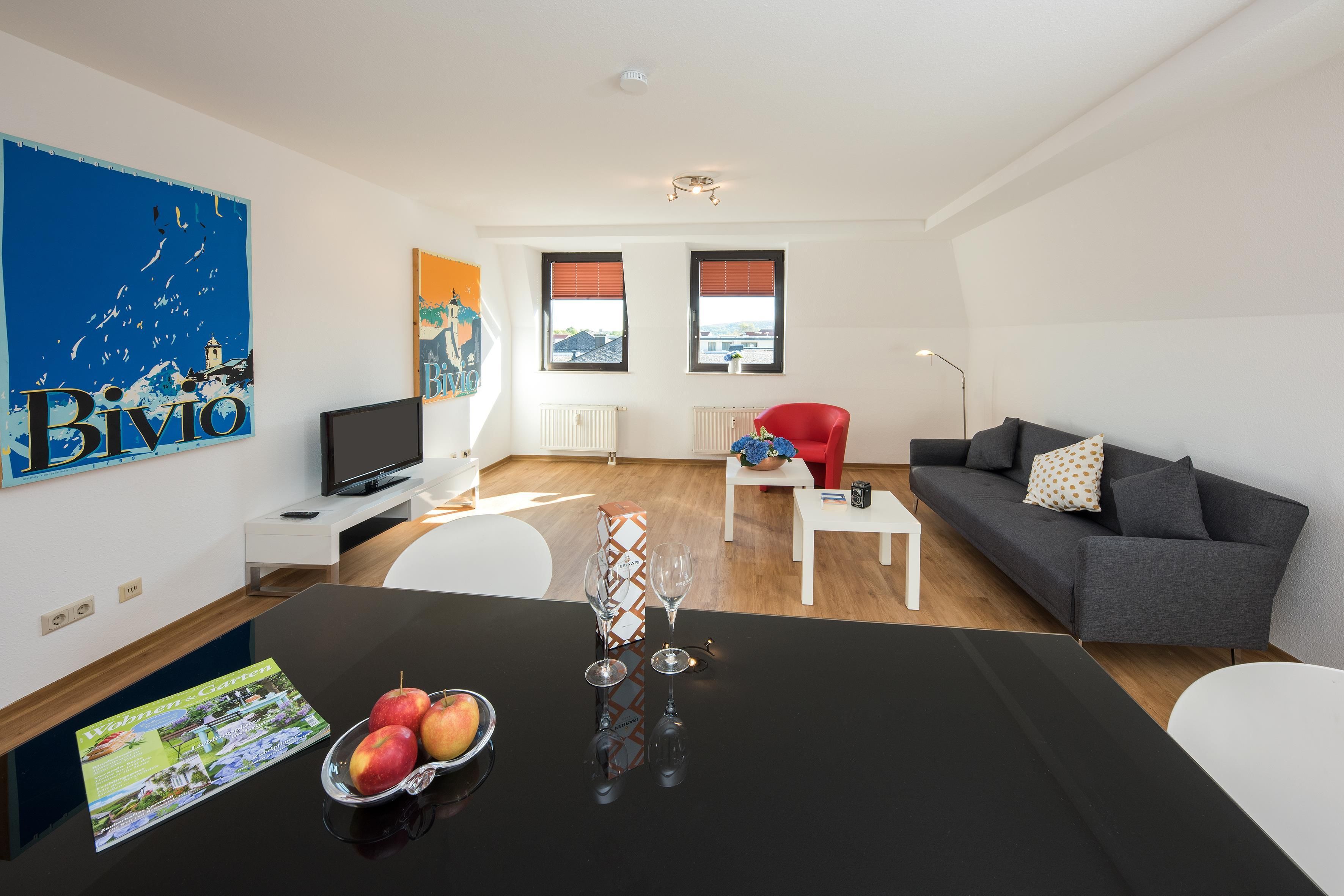 Blick über den schwarzen Glastisch mit Obstschale in den Wohnbereich eines Apartments der City Residenz mit grauem Sofa, rotem Sessel und Postern
