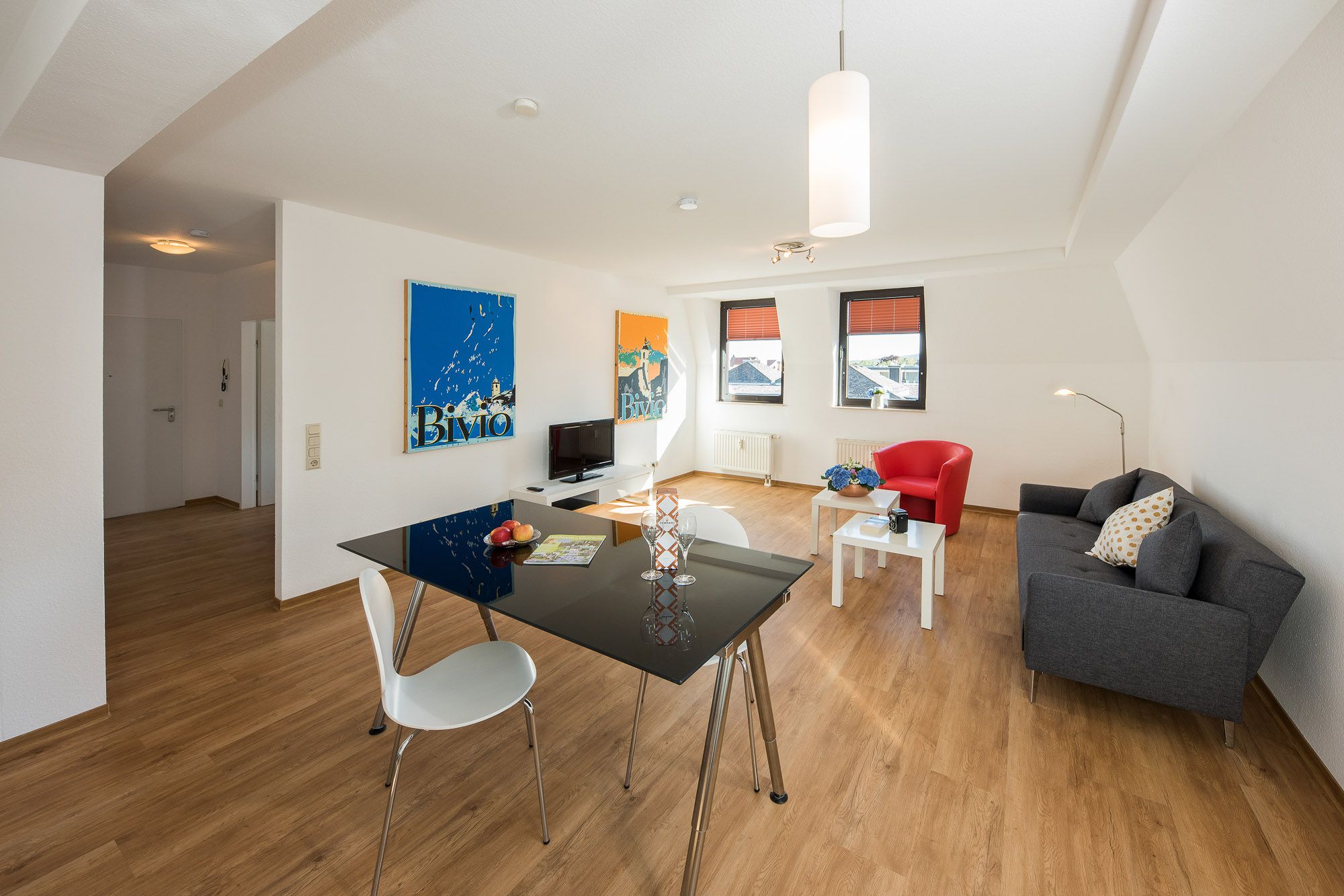 Apartment der City Residenz mit Glastisch, weißen Stühlen, grauem Sofa, rotem Sessel und Postern unter der Dachschräge