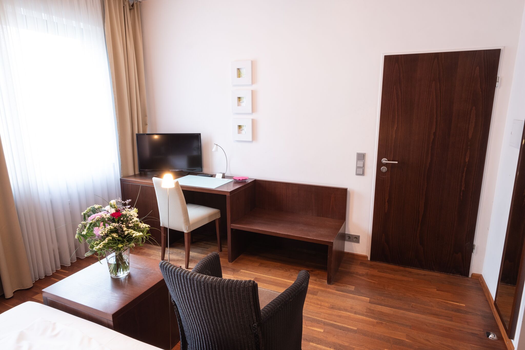 Studio der City Residenz mit Schreibtisch aus dunklem Holz, Fernseher, Korbstühlen und einem Blumenstrauß