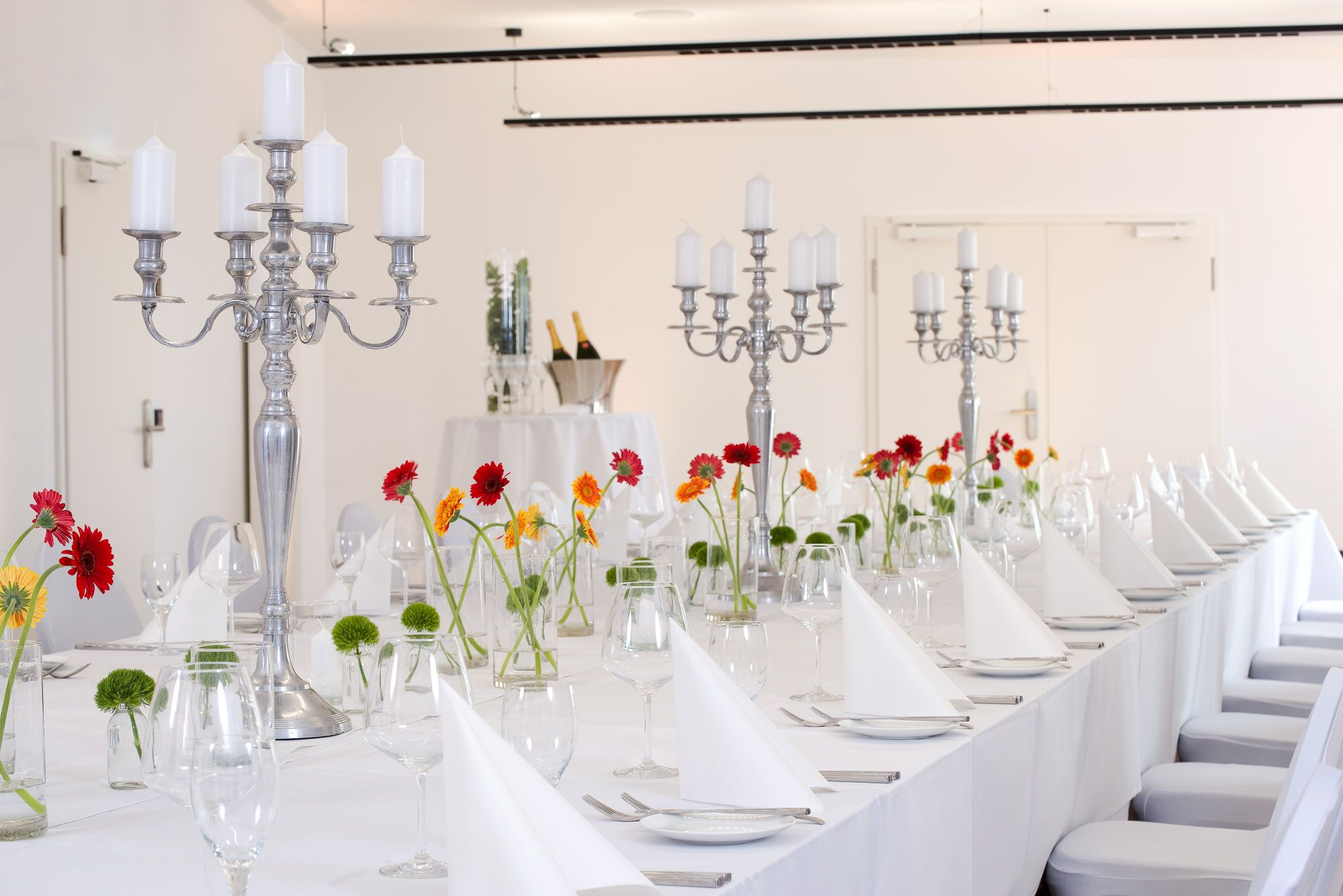 Lange weiß eingedeckte Festtafel mit silbernen Kerzenleuchtern, bunten Gerbera und Sektkühler im Hintergrund