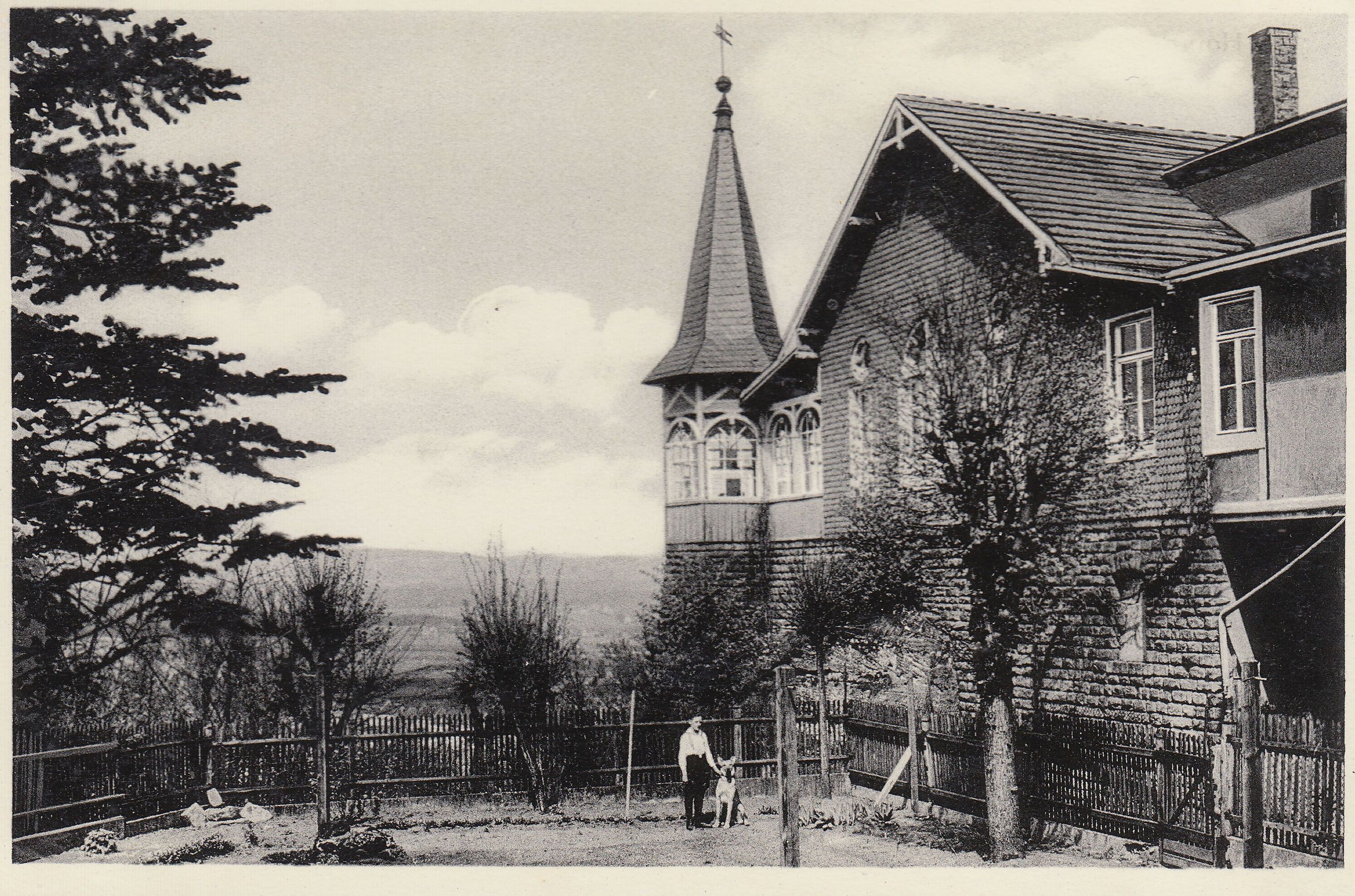 Historisches Schwarz-Weiß-Foto eines Hauses mit spitzem Türmchen, davor ein Mann mit Hund am Holzzaun