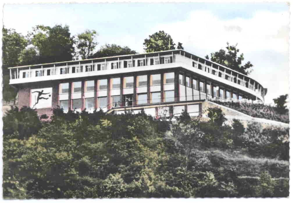 Kolorierte Postkarte eines langgestreckten Flachdachbaus am bewaldeten Hang, an der Fassade ein Fußballer-Relief