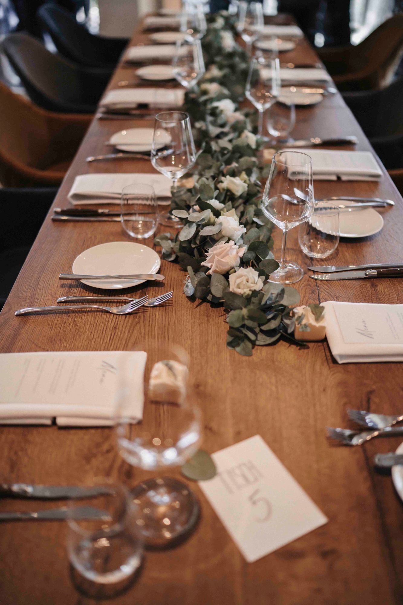 Lange Holztafel mit Eukalyptus-Girlande, weißen Blüten, Gläsern und handgeschriebenen Tischkarten