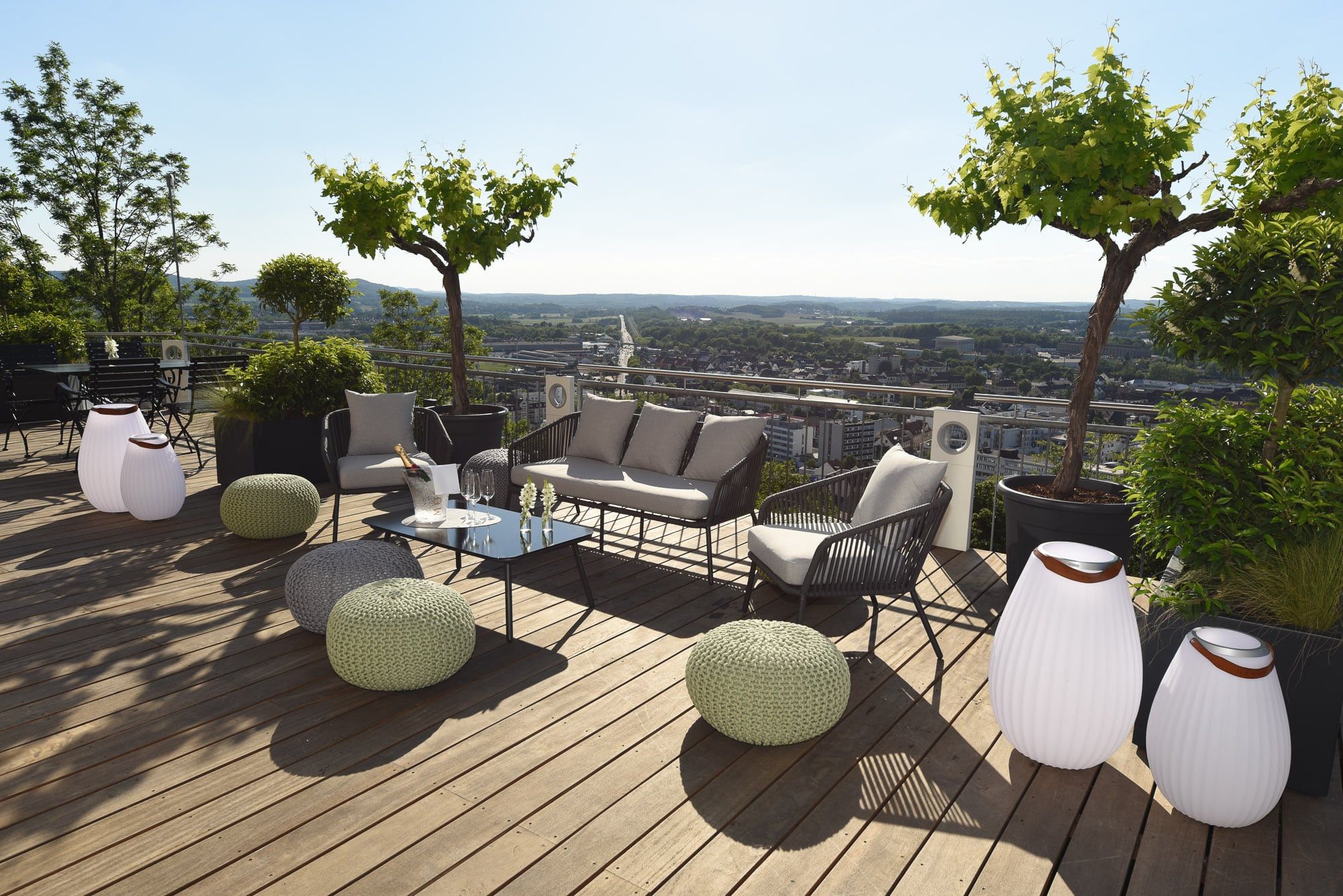 Lounge-Möbel, Strickhocker und Leuchtvasen auf der Holzterrasse, dahinter der Blick über die Dächer von Homburg