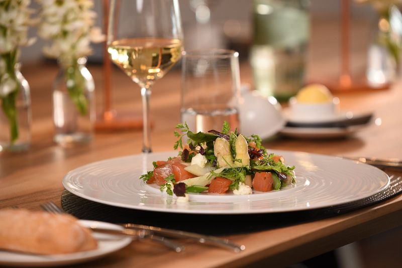 Salat mit Lachswürfeln, weißem Spargel und Kräutern auf weißem Teller, dahinter ein Glas Weißwein