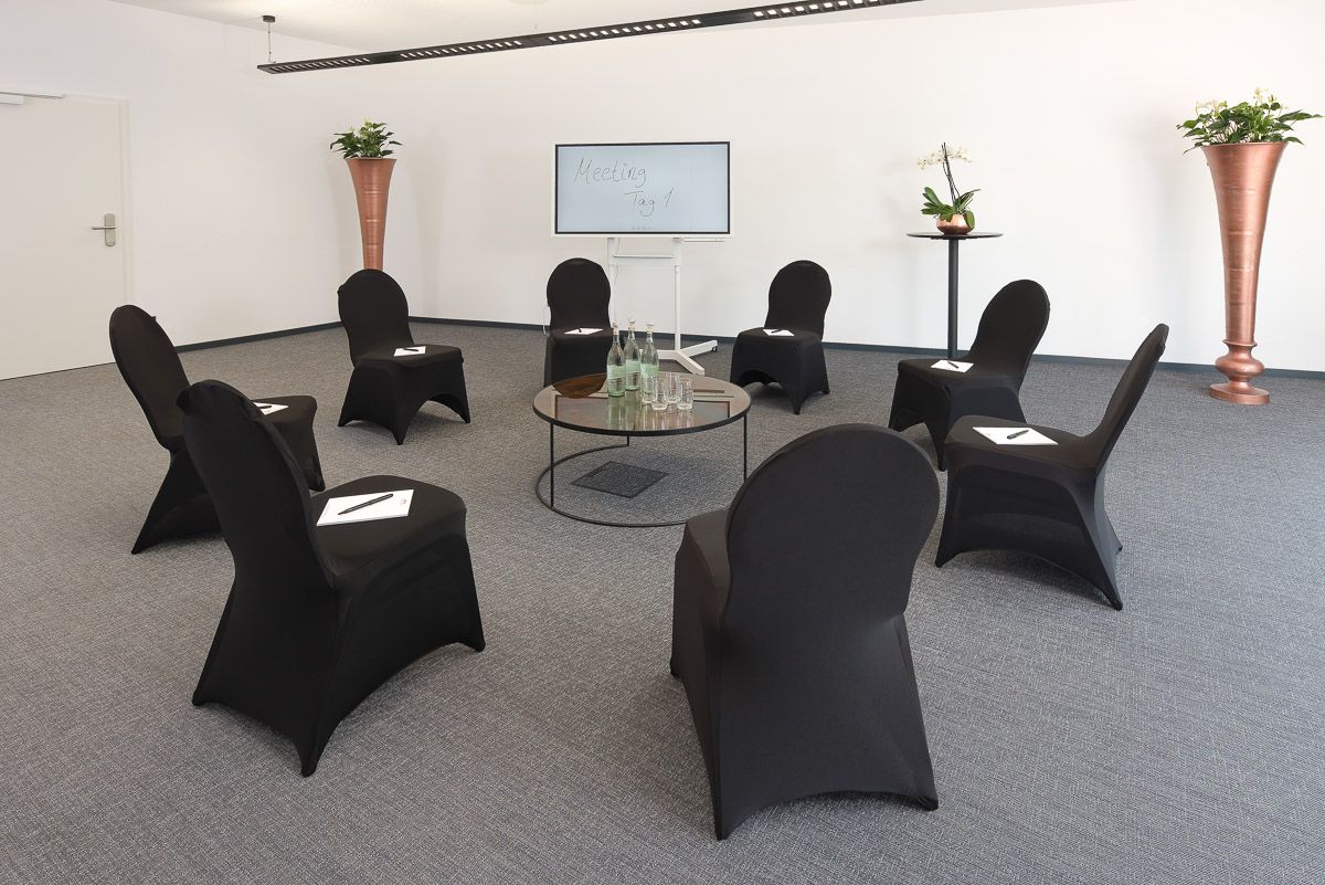 Stuhlkreis mit schwarz bezogenen Stühlen und Schreibblöcken, im Hintergrund ein Bildschirm mit „Meeting Tag 1“