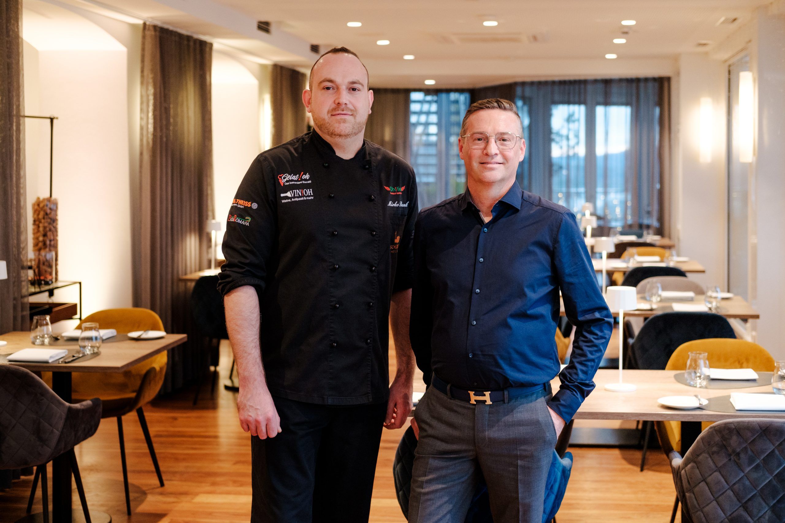 Koch in schwarzer Kochjacke und ein Mitarbeiter aus dem Service in dunkelblauem Hemd stehen nebeneinander im Restaurant, dahinter gedeckte Tische