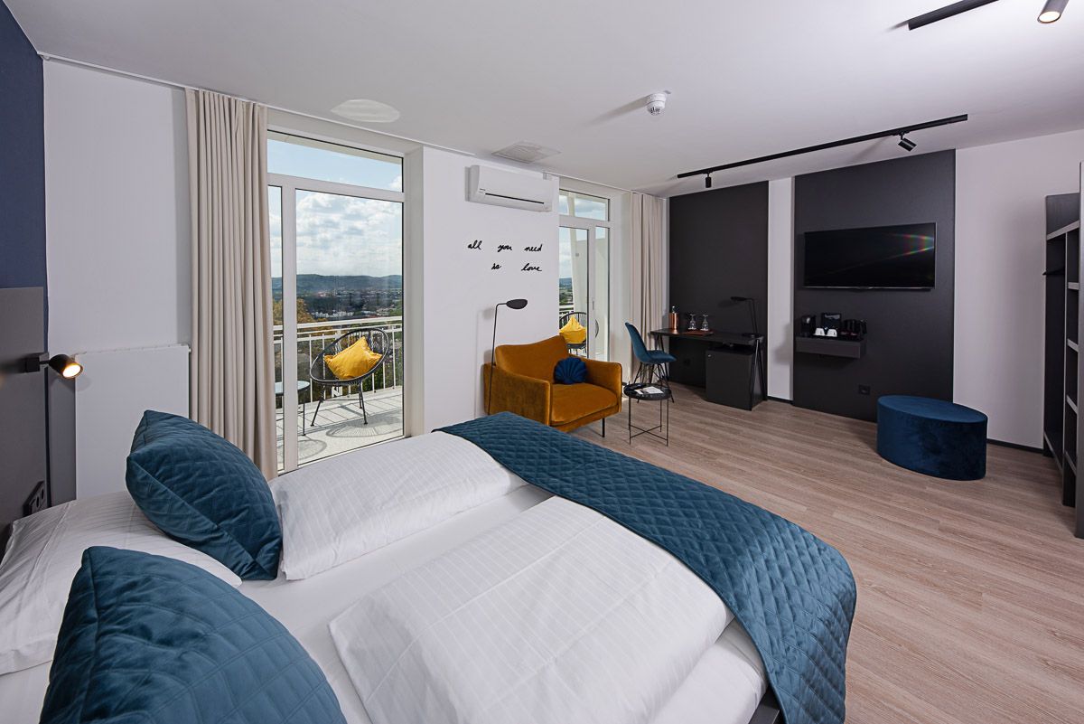 Blick vom Doppelbett zur offenen Balkontür, draußen ein Sessel mit gelbem Kissen und bewaldete Hügel über der Stadt