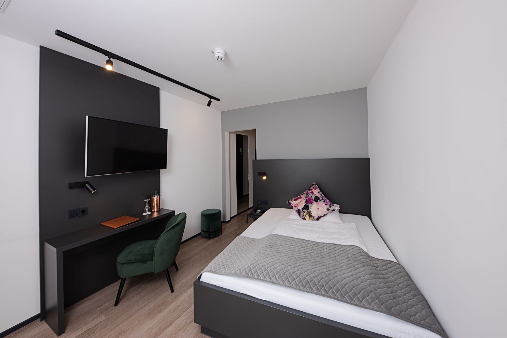 Business-Einzelzimmer mit Einzelbett, Schreibtisch mit grünem Samtstuhl und Wandfernseher auf dunkler Wandverkleidung