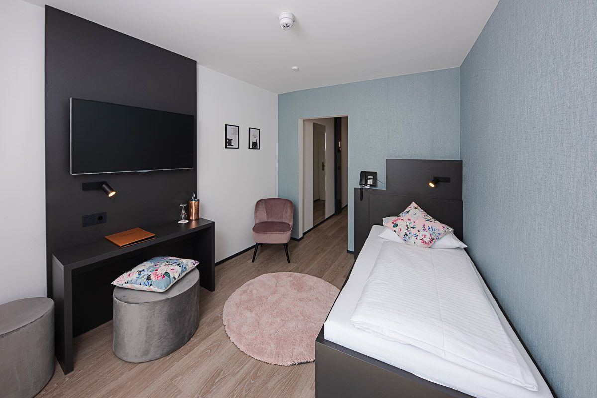 Comfort-Einzelzimmer mit Einzelbett an hellblauer Wand, Schreibtisch, rosa Sessel und rundem Hochflorteppich