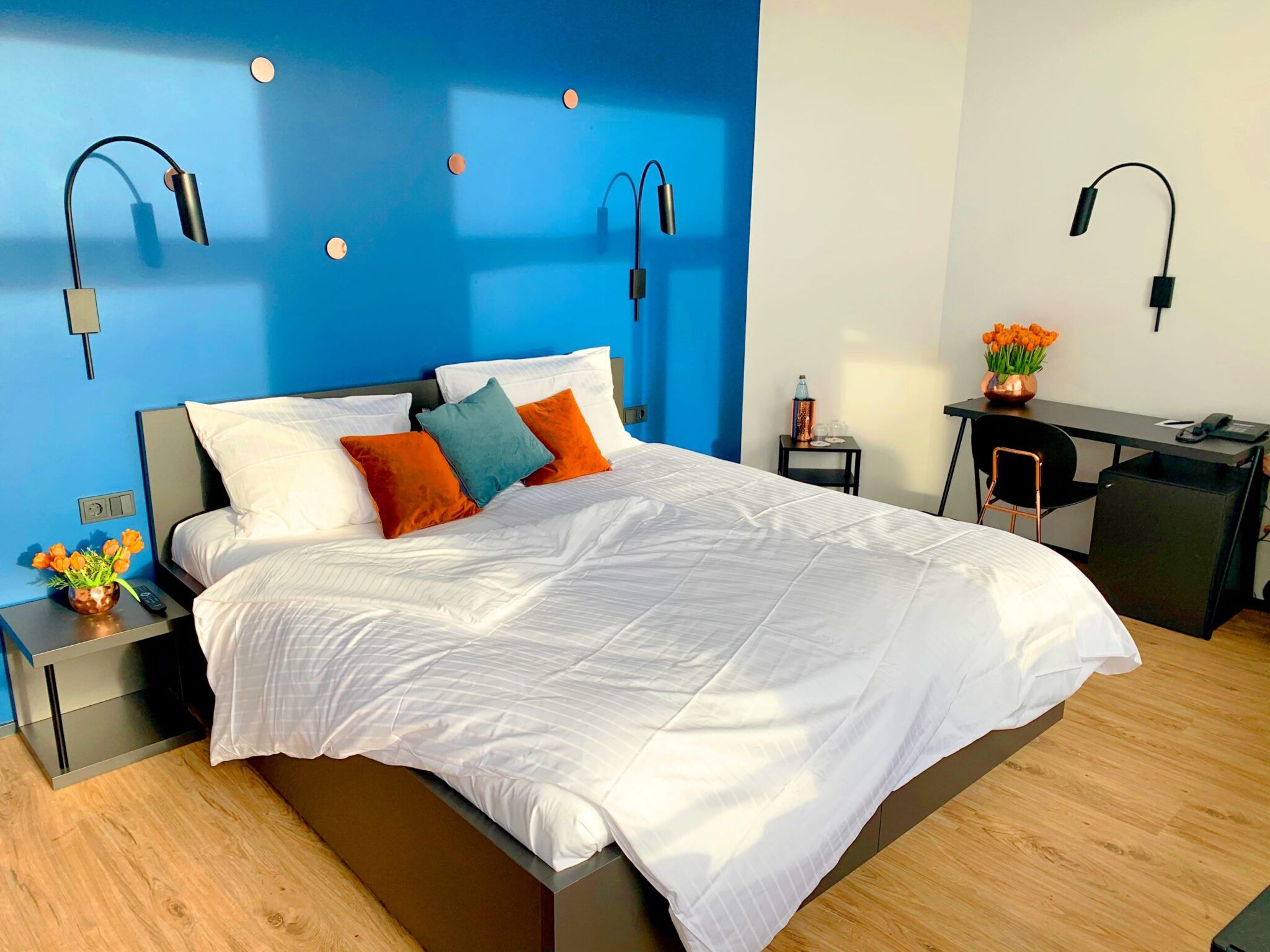 Deluxe-Zimmer mit Doppelbett vor kräftig blauer Wand, orangefarbenen Kissen und Tulpen auf Nachttisch und Schreibtisch