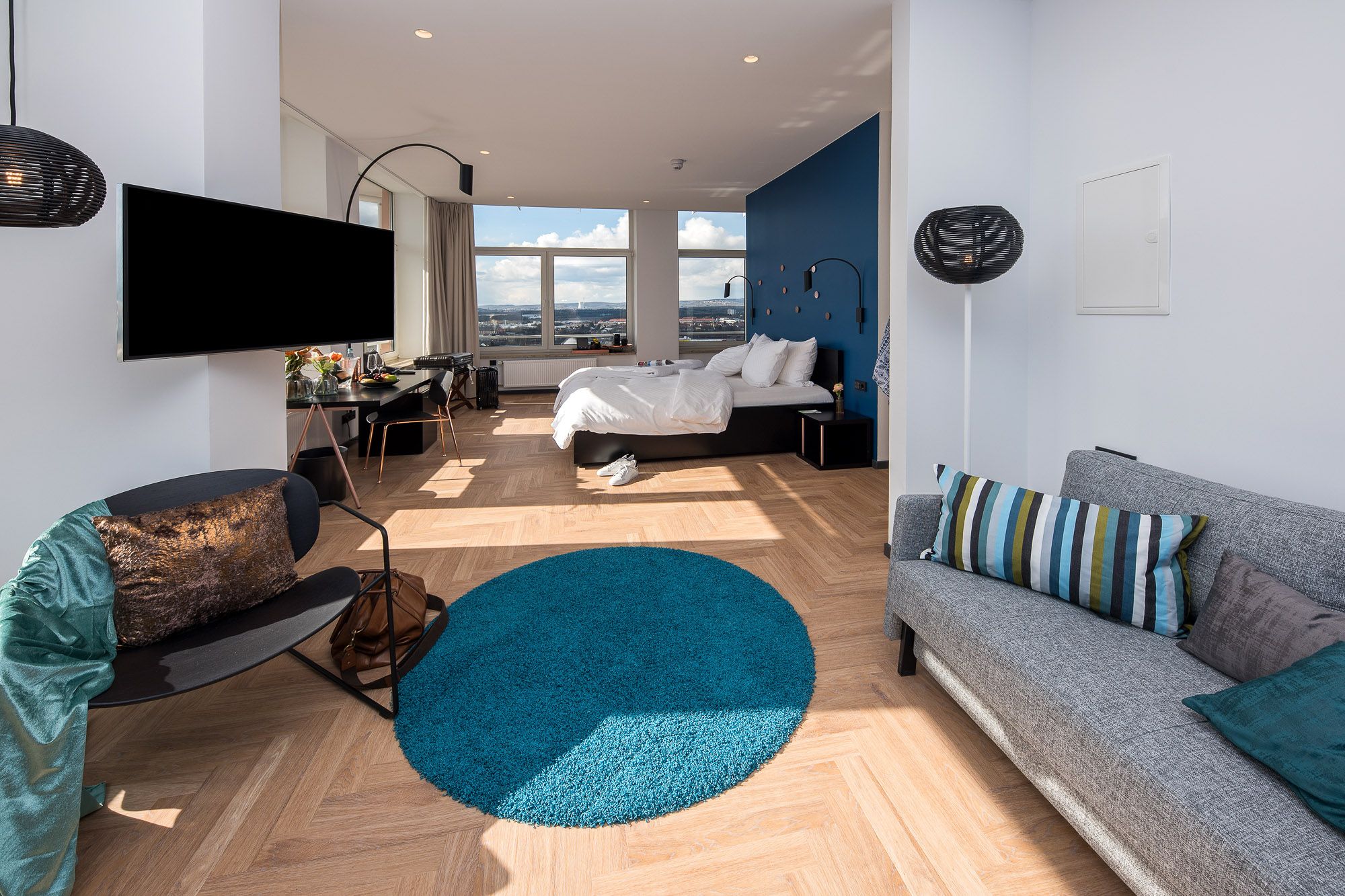 Suite mit Sofa, rundem petrolfarbenem Teppich und Fischgrätparkett, hinten das Bett vor breiten Fenstern